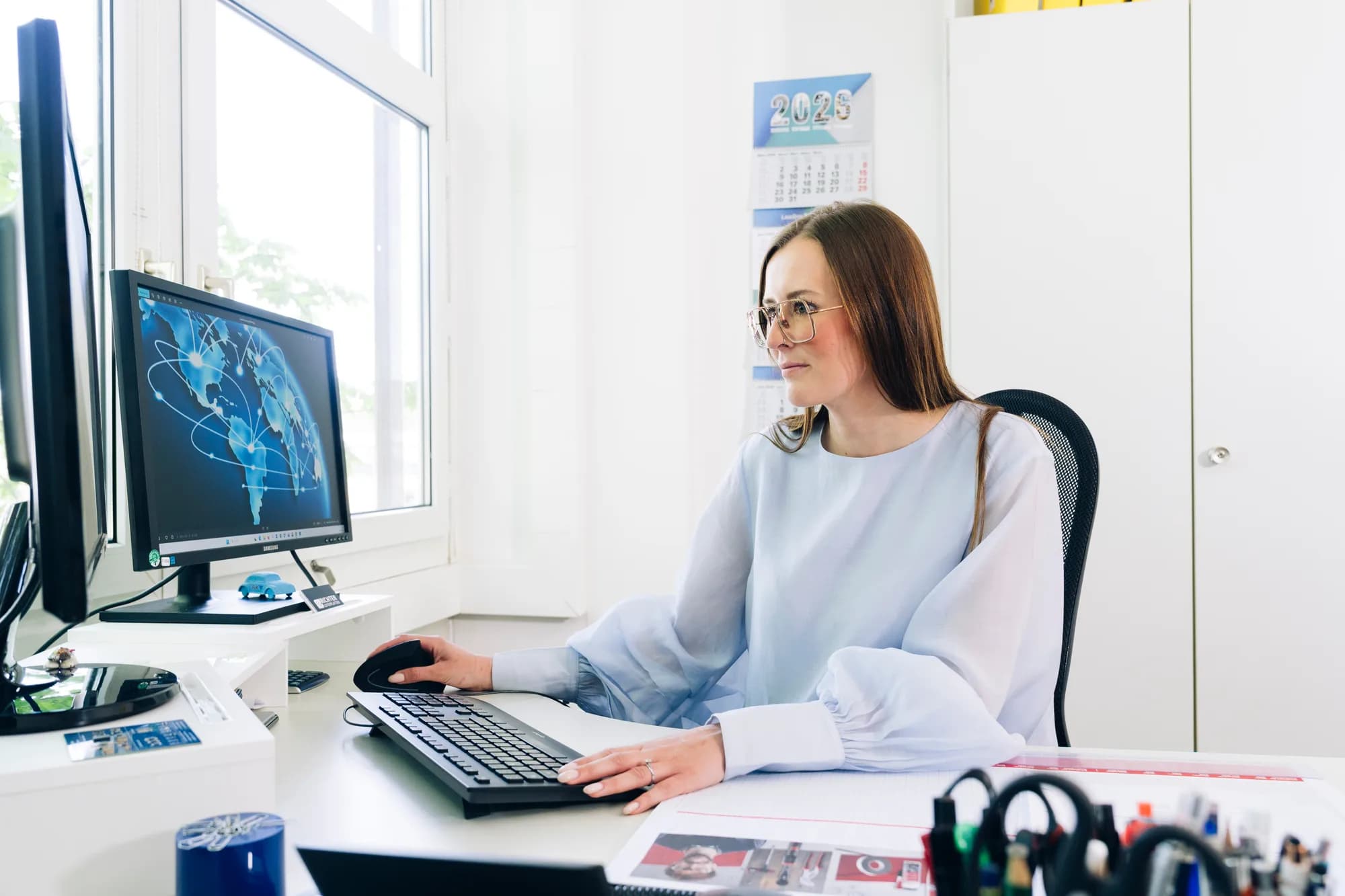 Konzentrierte Frau arbeitet an einem Schreibtisch in einem hellen Büro; Computermonitor, Tastatur, Dokumente und Kalender 2026 sind sichtbar.