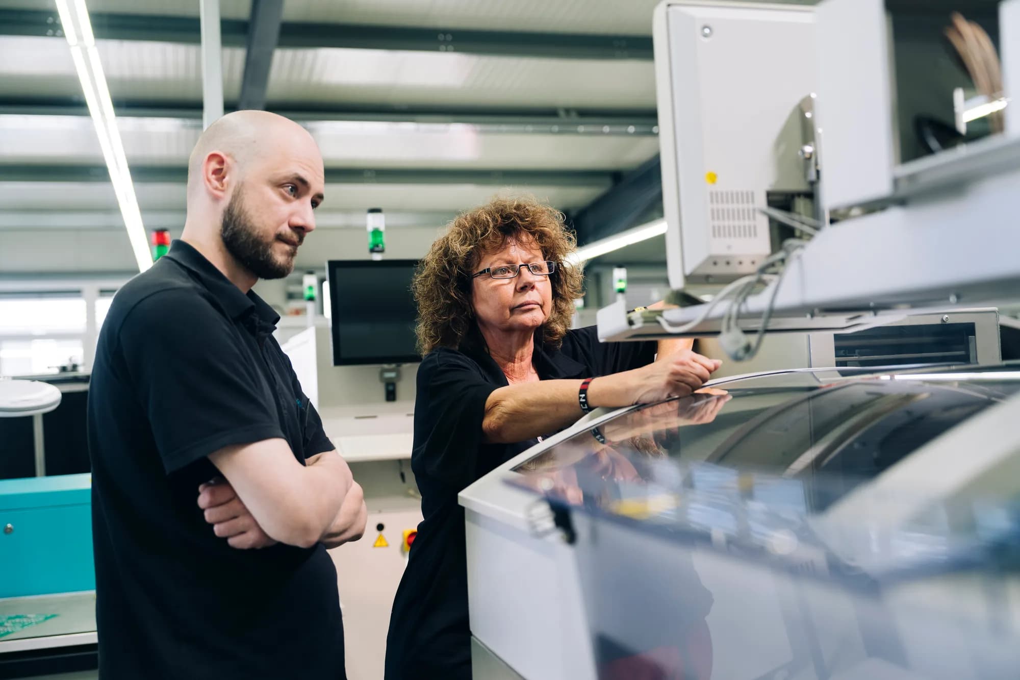 Mann und Frau in industrieller Elektronikwerkstatt. Vorne reflektierendes weißes Arbeitsgerät, Hintergrund mit elektronischen Komponenten und Regalen.
